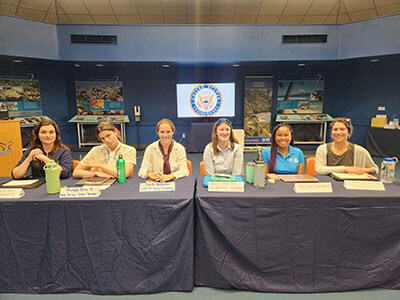a group of women sit at a table facing the camera with name tags in front of them