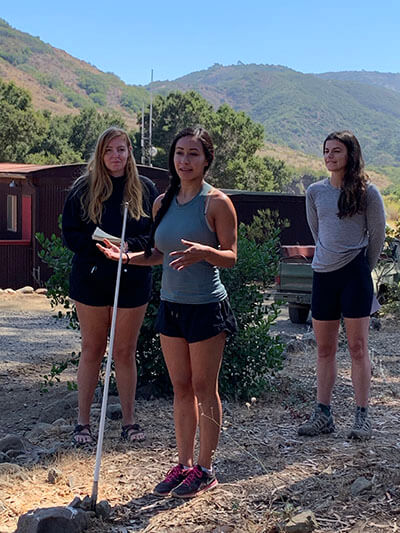 Three women stand with mountains in the background. The one in the middle speaks into a make-shift fake microphone